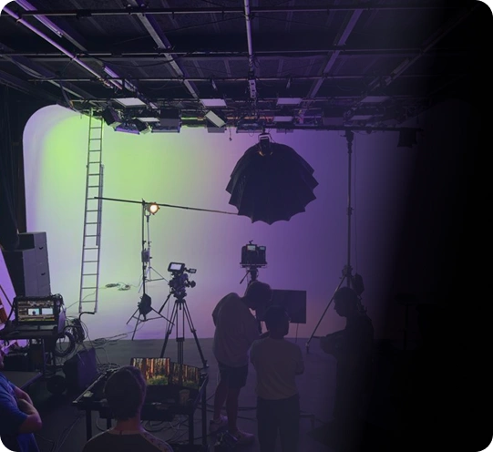 Studio filmowe z ekipą przygotowującą się do pracy. Jasno oświetlone tło z przejściami kolorów, widać kamery i sprzęt oświetleniowy. Zajęta, współpracująca atmosfera.