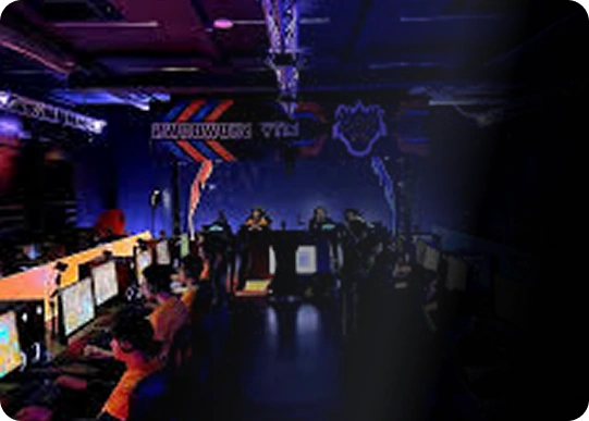 Słabo oświetlona arena gamingowa z neonowymi niebieskimi i pomarańczowymi światłami. Rzędy graczy przy stanowiskach komputerowych, skupieni na ekranach. Pełna energii, konkurencyjna atmosfera.