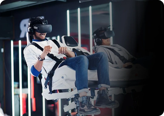 Dwie osoby w zestawach VR siedzą na fotelach symulatora ruchu, wyrażając ekscytację. Otaczają je neonowe światła, sugerujące środowisko gamingowe lub technologiczne.